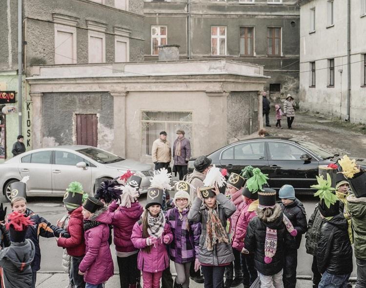 Zdjęcie przedstawiające grupę dzieci w kolorowych ubraniach i czapkach górników, posród szarych, betonowych budynków
