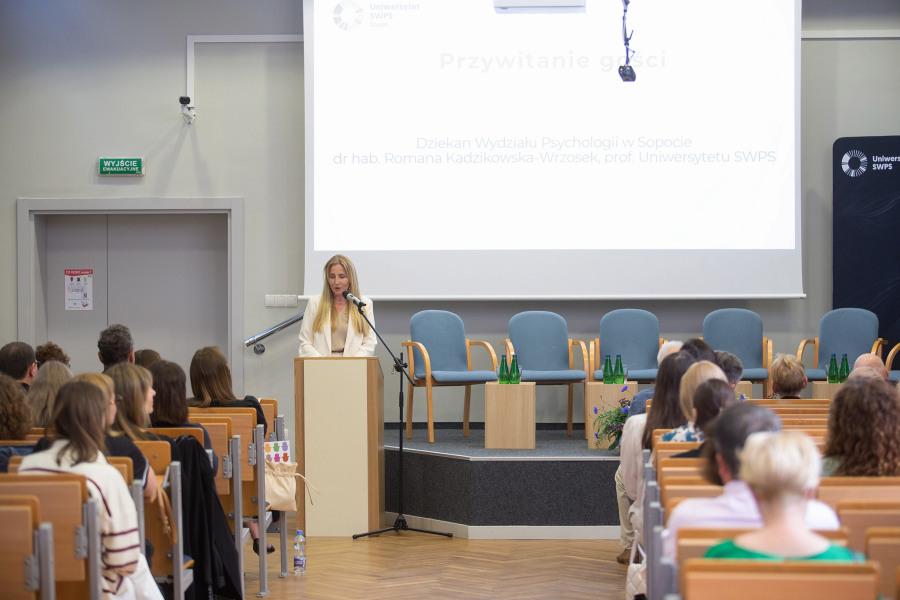 Dziekan Wydziału Psychologii w Sopocie, prof. Romana Kadzikowska-Wrzosek, stoi na mównicy i wita zebranych gości, siedzących na krzesłach