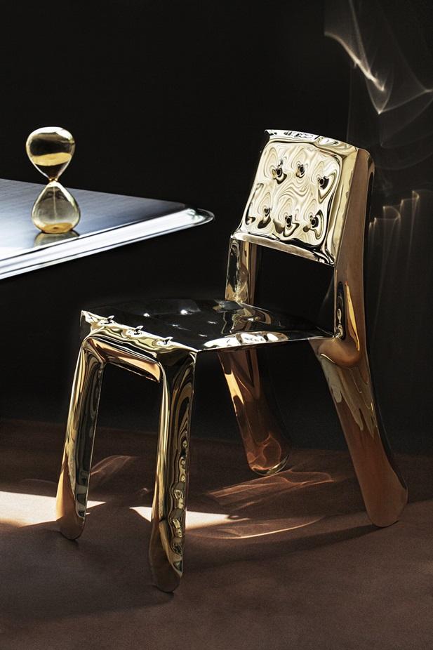 Złote krzesło wykonanie z dmuchanego metalu stojące obok stołu, na którym znajduje się klepsydra odmierzająca czas.