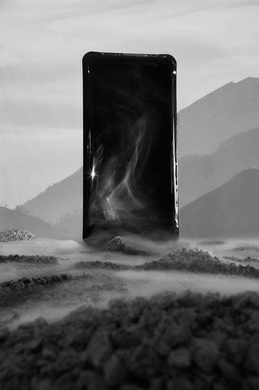 Prostokątny fragment nadmuchanego metalu, przypominający kształtem ekran smartfona. Stoi na kamienistej powierzchni, jest otoczony delikatną mgłą i oparami.