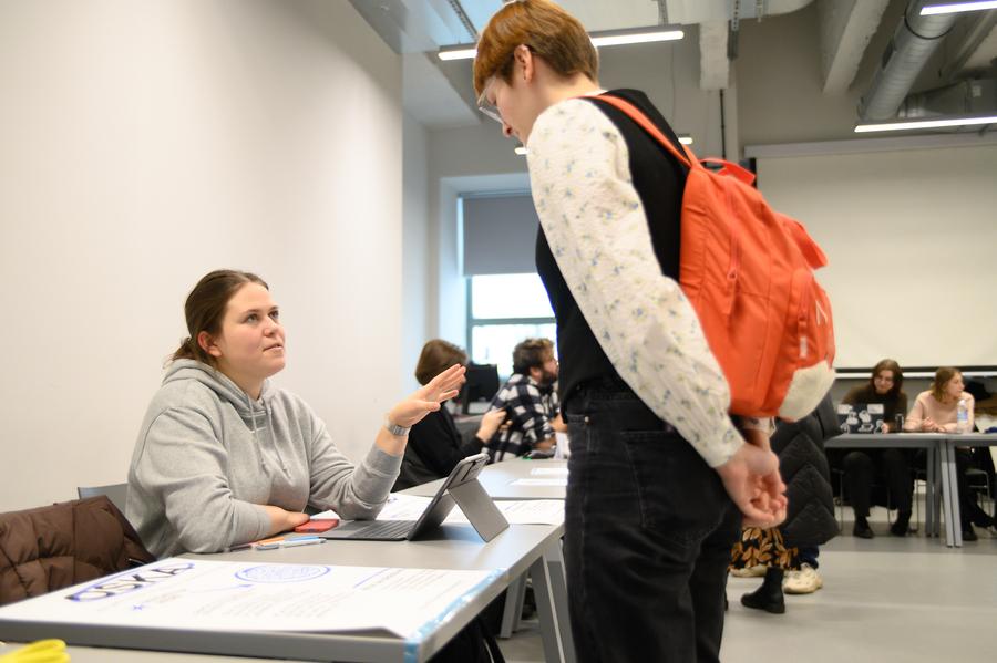 Studentka stoi przy stoisku OSKA, a siedząca przy biurku członkini tej organizacji coś jej tłumaczy, gestykulując