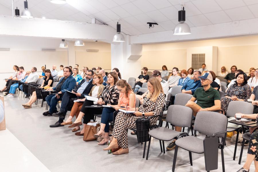 Dużo ludzi siedzi na krzesłach. Część osób robi notatki