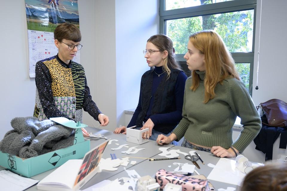 Agata Nowak rozmawiająca z dwoma studentkami przy stole, na którym leżą przybory do pracy i kartki papieru