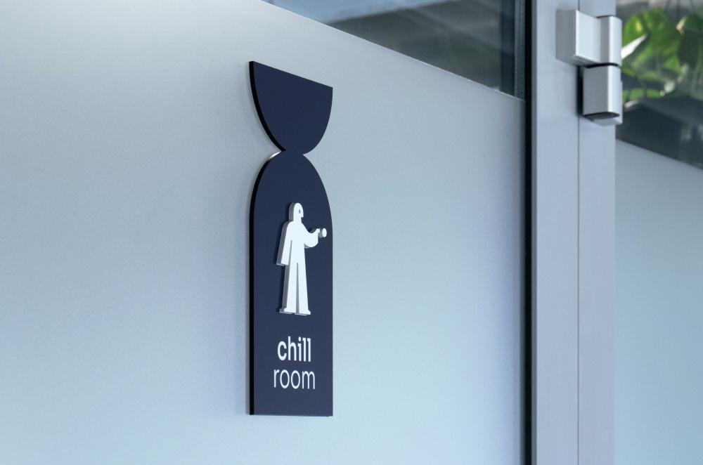 Ciemnoniebieska tabliczka informacyjna na matowej szybie przedstawiająca biały piktogram stojącej postaci oraz napis „chill room”.