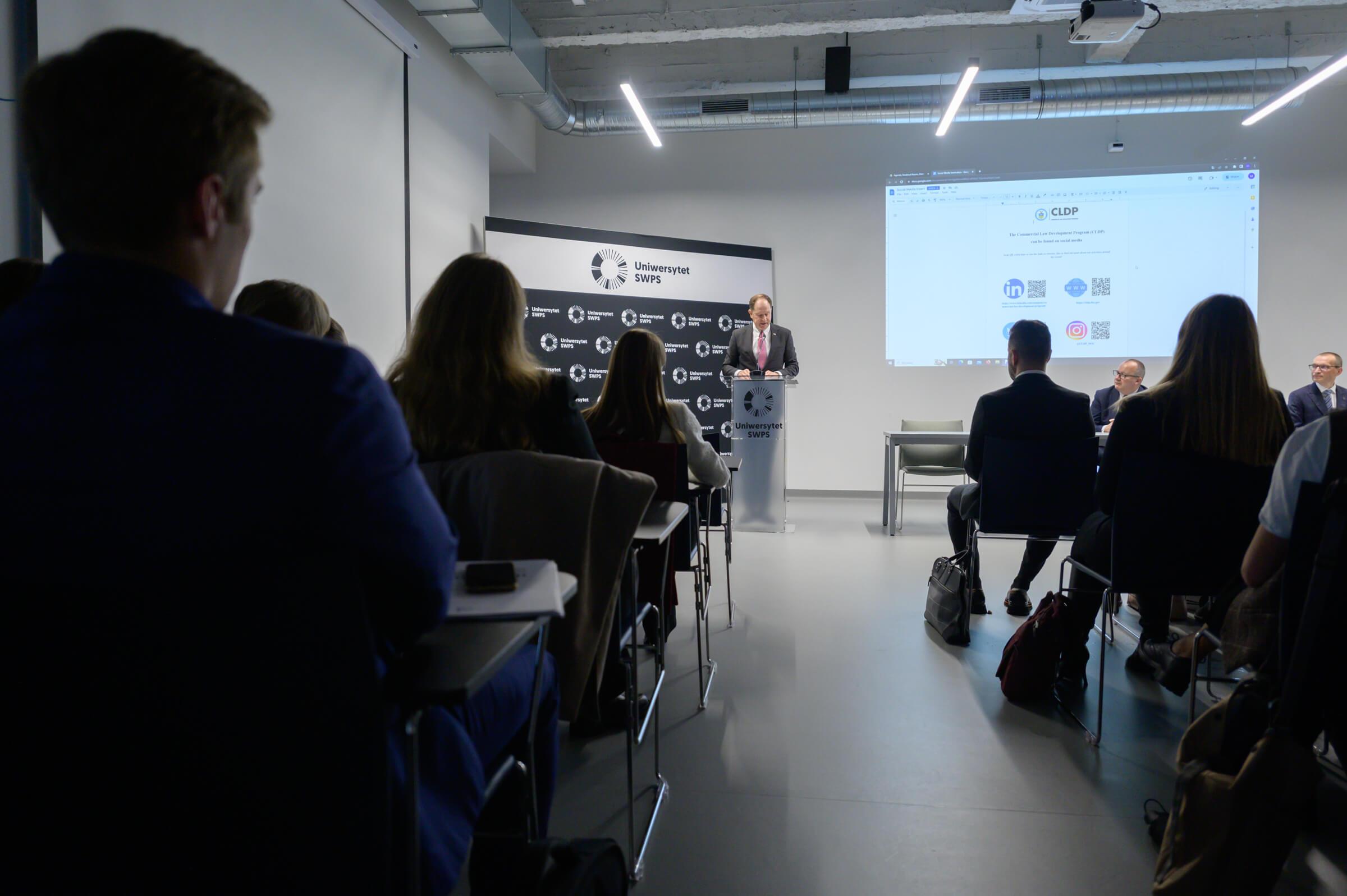 Widok z tyłu na publiczność podczas otwarcia szkolenia Vis Moot Bootcamp na Uniwersytecie SWPS w Warszawie, w tle przemawia ambasador USA, Mark Brzeziński