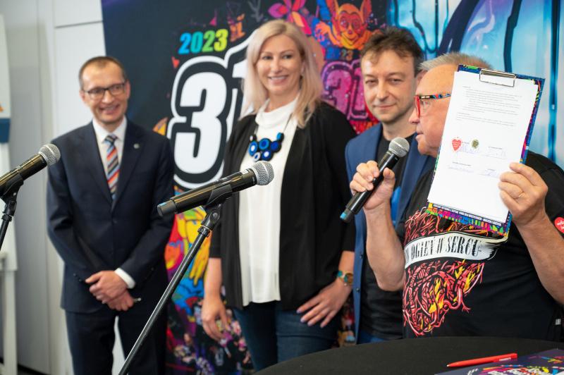 Jerzy Owsiak prezentuje publiczności podpisany list intencyjny. W tle uśmiechnięci prof. Roman Cieślak, rektor Uniwersytetu SWPS, Dominika Kacperek-Karbowiak, dyrektor zarządczy Kliniki Uniwersytetu SWPS, oraz Piotr Ciszek, prezes RESQL sp. z o.o.