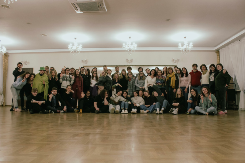 Zdjęcie grupowe około 50 chórzystek i chórzystów zrobione podczas wyjazdowych warsztatów chóralnych. Studentki i studenci pozują do zdjęcia w codziennych ubraniach. Uśmiechają się