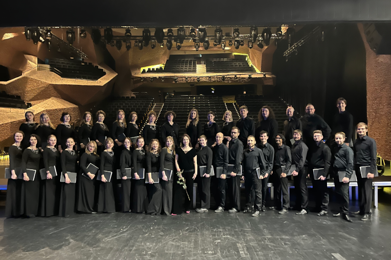 Centrum Kulturalno-Kongresowe Jordanki w Toruniu. Na scenie stoją chórzyści i chórzystki w eleganckich czarnych strojach chóralnych, trzymając zeszyty z nutami z czarnymi okładkami. Na środku zdjęcia stoi dyrygentka chóru, Ewa Mackiewicz. Trzyma w prawej ręce różę. Nad sceną wisi rozbudowany, profesjonalny sprzęt oświetleniowy