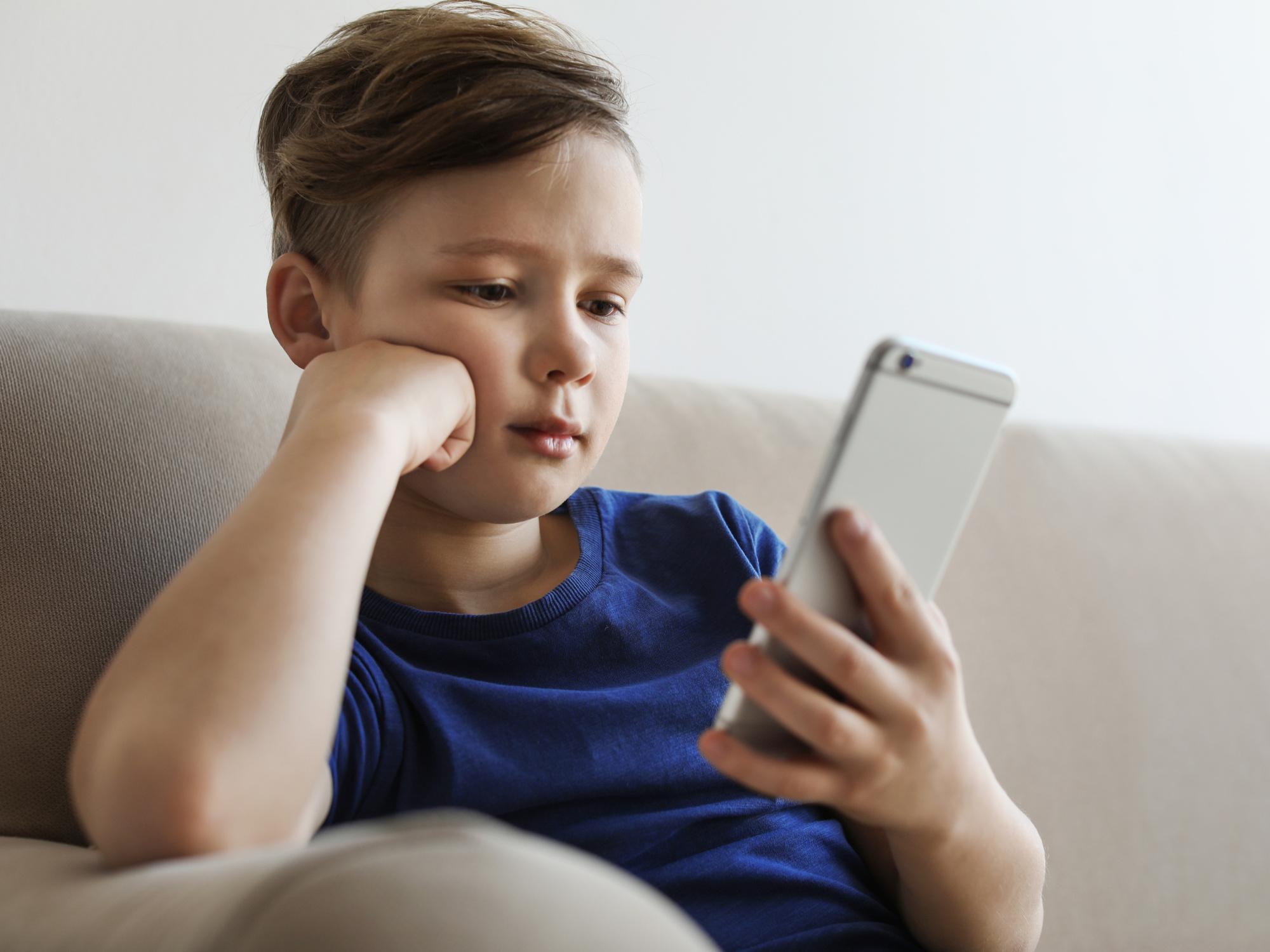 Chłopiec siedzi na kanapie i wpatruje się w ekran smartfona, podpierając głowę dłonią