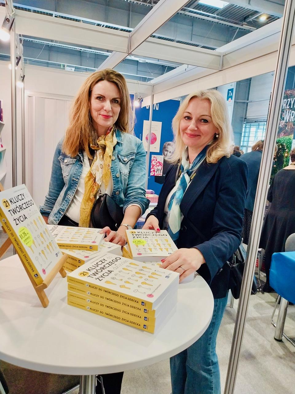 Doktor Joanna Kwaśniewska i Dorota Siudowska-Mieszkowska stoją przy okrągłym stoliku, na którym leży kilkanaście egzemplarzy książki „21 kluczy do twórczego życia dziecka”