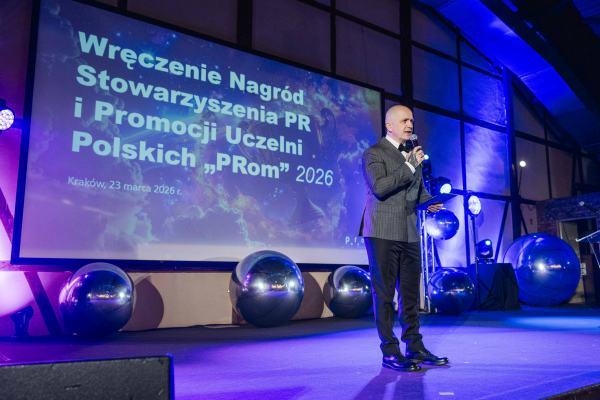 Gala Nagrody Stowarzyszenia PRom 2026.  Mężczyzna w garniturze przemawia do mikrofonu. W tle slajd z informacją: „Wręczenie Nagród Stowarzyszenia PR i Promocji Uczelni Polskich „PRom” 2026