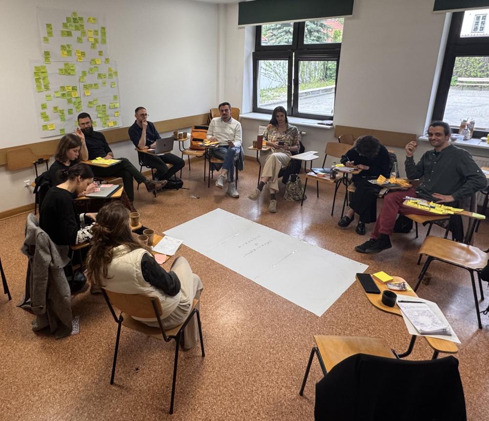 Grupa ekspertek i ekspertów siedzi na krzesłach ustawionych w kółku, na podłodze między nimi leży duża kartka. Na ścianie w tle wisi kartka z mnóstwem zapisanych karteczek samoprzylepnych