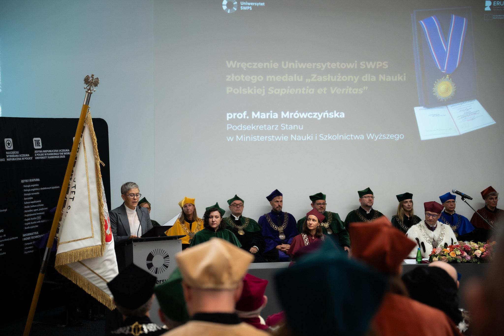 Fotografia przedstawia prof. Marię Mrówczyńską przemawiającą z mównicy, za którą siedzą inni profesorowie w strojach akademickich w auli Uniwersytetu SWPS. W tle duży ekran wyświetlający informacje o wydarzeniu oraz zdjęcie medalu i listu gratulacyjnego. Na ekranie widać logotypy USWPS oraz Ministerstwa Nauki i Szkolnictwa Wyższego.