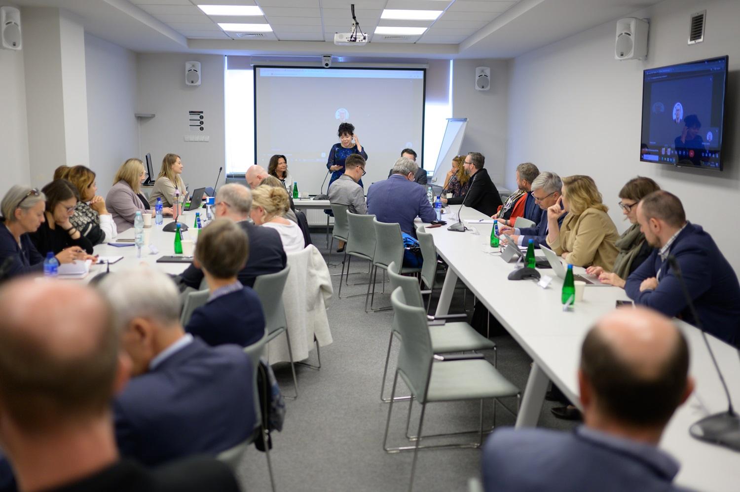 zdjęcie sali, w której odbywa się konferecja. Widać gości i gościnie siedzących przy stołach, a tle siedzą prowadzace spotkanie