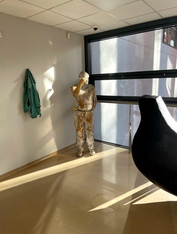 Rzeźba mężczyzny w średnim wieku, w czapce i ubraniach roboczych, trzymający w kieszeni banana. Na ścianie wisi powieszona na wieszaku kurtka.