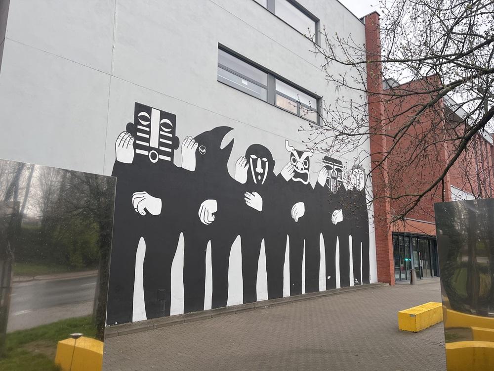 Mural przedstawiający sześć postaci ustawionych w jednym rzędzie. Pięć z nich nosi maski. Twarz szóstej postaci nie jest zakryta maską i nadstawia ucho w kierunku pozostałych.