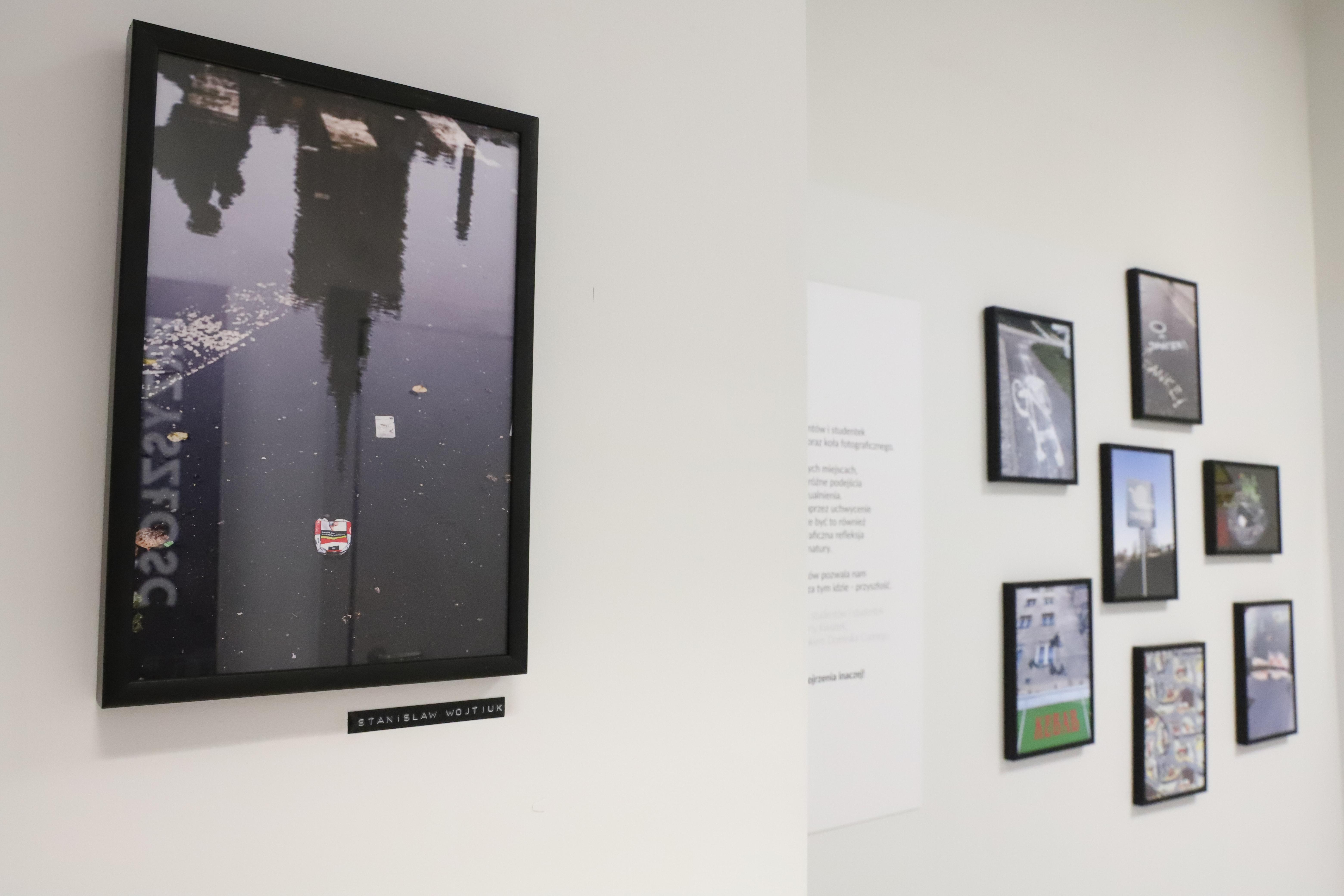 Zdjęcie 7 prac artystów w czarnych ramkach wyeksponowanych na ścianie. Praca na pierwszym planie przedstawia odbicie pałacu kultury na mokrym od deszczu chodniku.