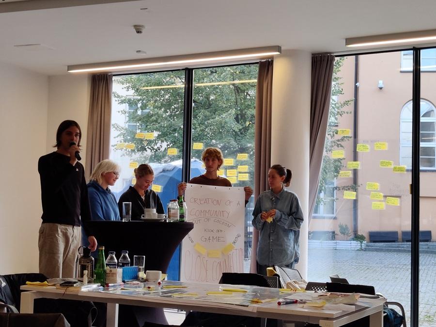 Grupa uczestników warsztatów prezentująca swój projekt. Przewodniczący grupy mówi coś przez mikrofon, stojący obok uczestnik trzyba plakat, na którym rozpisana jest nazwa i koncepcje projektu. Na szybie zanimi widać dużo karteczek samoprzylepnych.